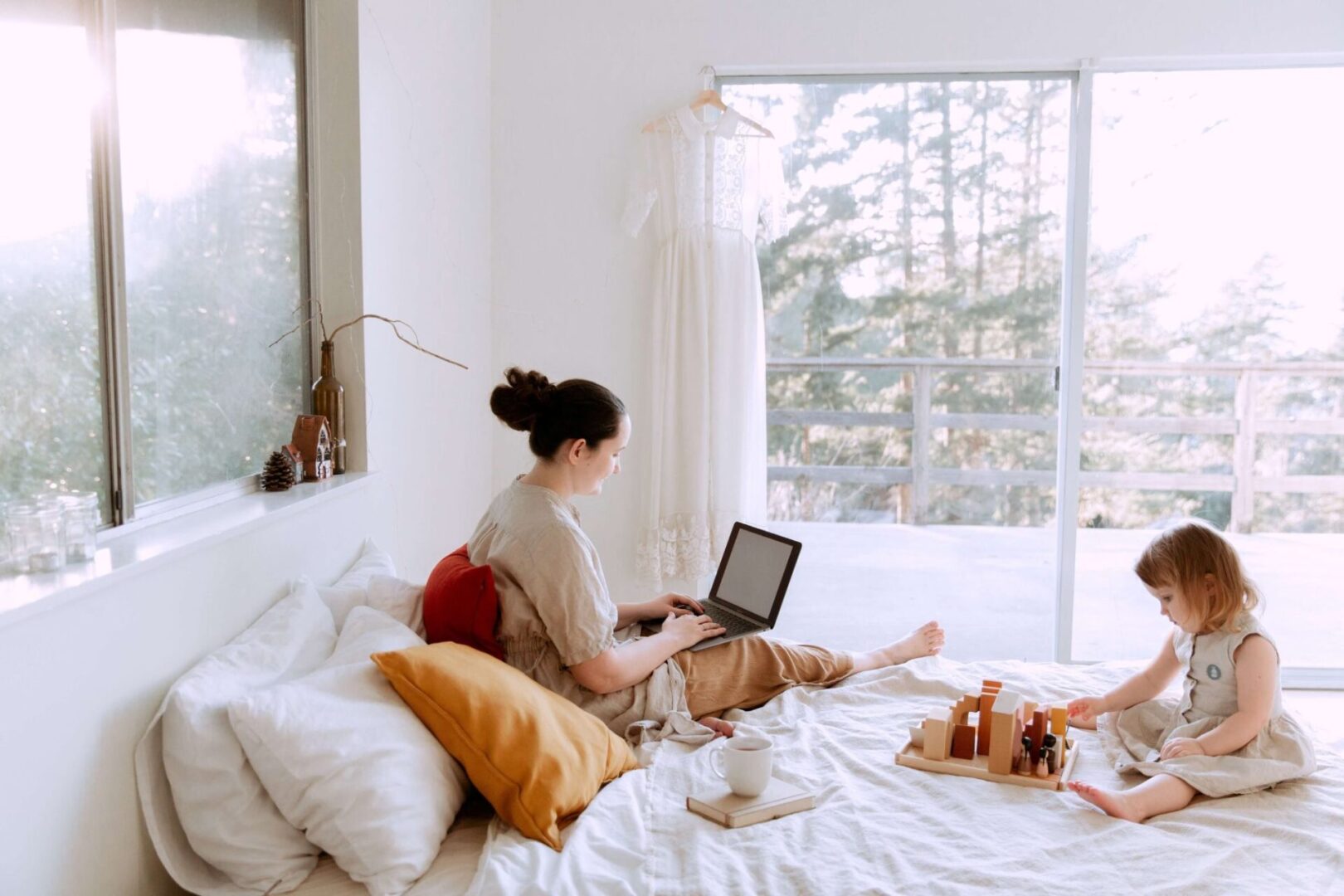 Girl sitting on bed and playing with wooden blocks and toys while mother using laptop